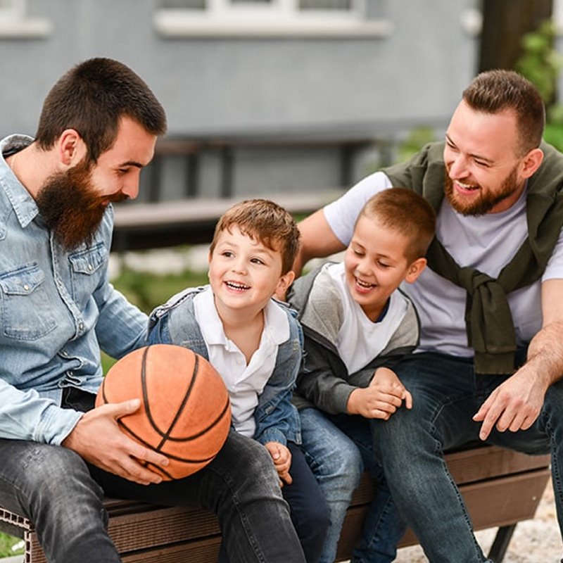 two dads and their two sons sitting on a bench outside with a basketball